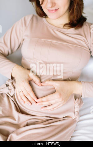 Les femmes enceintes smiling brunette dans une robe beige mit sa main sur son ventre pour se sentir en forme de coeur à l'amour du bébé. concept de l'amour, la grossesse. Banque D'Images