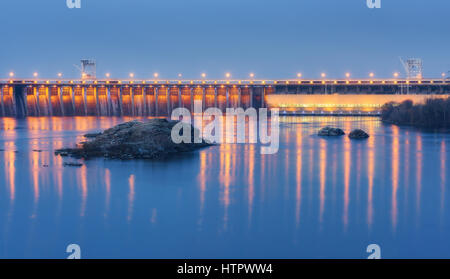 Barrage de nuit. Beau paysage industriel avec station d'énergie hydroélectrique du barrage, pont, rivière, illumination de ville reflète dans l'eau, les roches et le ciel. Banque D'Images