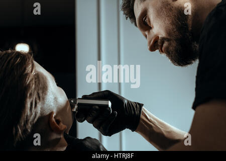 Thème BARBERSHOP. Coiffure BARBU EN NOIR GANTS DE CAOUTCHOUC EST LA BARBE DE FRAISAGE DE SON JEUNE BEAU CLIENT. Il est à l'aide d'une tondeuse à cheveux Banque D'Images