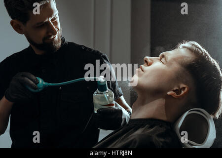 Thème BARBERSHOP. Coiffure BARBUS DANS DES GANTS EN CAOUTCHOUC NOIR LOTION DE RASAGE APRÈS LES PROJECTIONS DE LA BOUTEILLE DE JET SUR LA PEAU DU CLIENT Banque D'Images