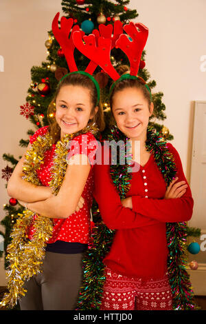 Twin Sisters posant devant un arbre de Noël avec des guirlandes autour du cou. Ils sont regardant la caméra et sourit. Banque D'Images