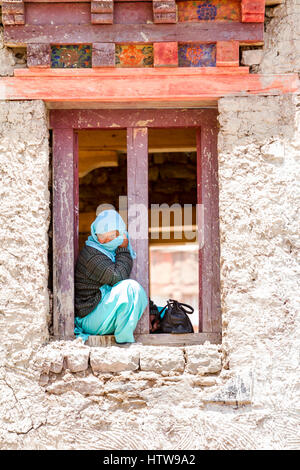 Hemis, Inde - le 29 juin 2012 : Lonely couverts avec châle young woman sitting in a windows span regardant un masqué et religieuses Cham en costume Dance Fest Banque D'Images