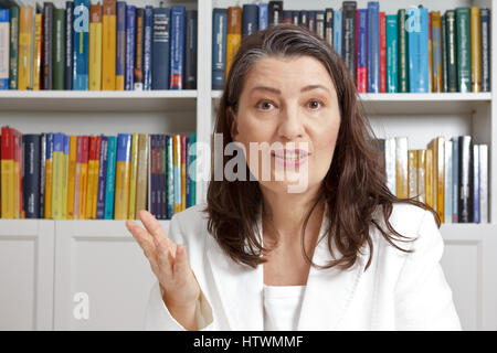 Femme d'âge moyen en blanc blazer expliquant quelque chose en face de livres dans une bibliothèque, enseignant, tuteur, conseiller, conseiller, psychothérapeute Banque D'Images