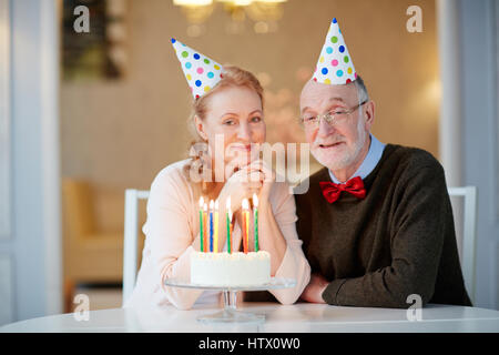 Portrait de senior couple celebrating birthday ensemble assis à table avec gâteau et wearing party hats smiling and looking at camera Banque D'Images
