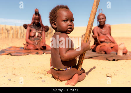 Jeune garçon enfant Himba est assis en face de deux femmes Himba dans un village dans le Nord de la Namibie. Banque D'Images