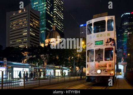 Un tramway de Hong Kong voyageant le long d'une rue dans le quartier des affaires avec des gratte-ciel en arrière-plan dans la nuit. Banque D'Images
