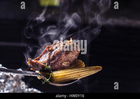 Smokeyshaesya la viande sur une cuillère, avec des épis de maïs et le poivre-2. Fond noir. Banque D'Images