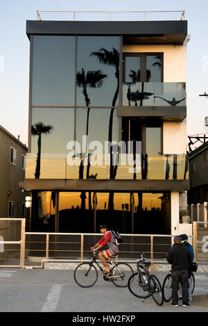 Un cycliste passe devant une maison de plage reflétant les palmiers et le soleil couchant à Venice Beach, Californie Banque D'Images