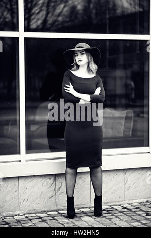 Portrait de jeune fille aux cheveux rouges de mode sur red hat et robe noire avec des composent posés contre la grande fenêtre. Photo en noir et blanc. Banque D'Images