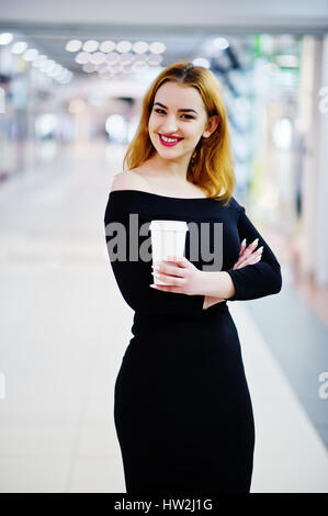 Fille aux cheveux rouges de mode de l'usure sur robe noire avec des faire holding Coffee cup au centre commercial du commerce. Style musclé photo filtres Instagram. Banque D'Images