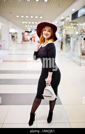 Fille aux cheveux rouges de mode de l'usure sur robe noire et red hat avec mesdames sac à dos posé au centre commercial du commerce. Style musclé photo filtres Instagram. Banque D'Images