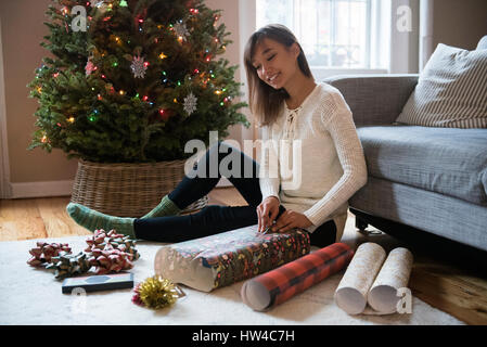 Mixed Race woman wrapping gift sur le plancher près de l'arbre de Noël Banque D'Images