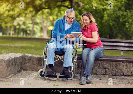 Heureux l'homme en fauteuil roulant avec l'aide de tablet fille dans le parc Banque D'Images