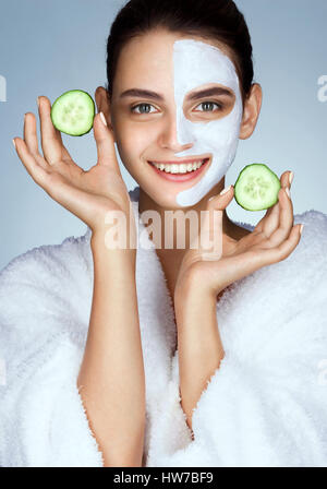 Rire beau modèle avec les tranches de concombre dans les mains. Photo de fille en peignoir blanc et soin du visage avec masque. Le toilettage lui-même Banque D'Images