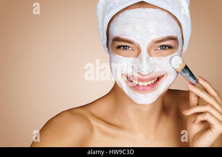 Belle jeune fille recevant le masque facial cosmétique. Portrait of smiling girl avec une serviette sur sa tête sur fond beige. Le toilettage lui-même Banque D'Images