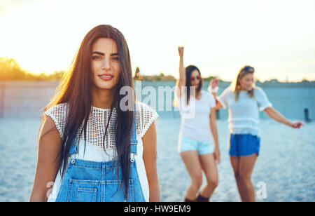 Calme belle jeune femme adulte dans la région de long cheveux brun et bleu jean bretelles sur plage avec deux amis danse Banque D'Images