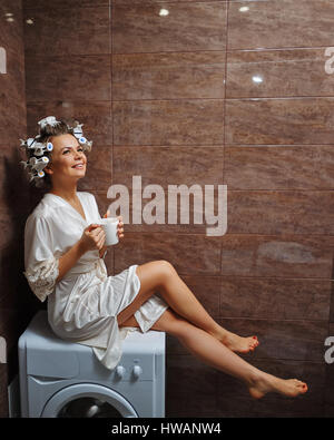 Jeune femme au foyer attrayant dans la salle de bains. Fille dans un peignoir et bigoudis boire le café du matin. Bonjour. Banque D'Images