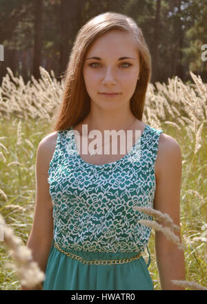 Belle jeune fille aux cheveux longs, vêtu de bleu, vert à l'extérieur, debout au milieu de la région ensoleillée de compensation pleine de fleurs, des oreilles, du blé en forêt Banque D'Images