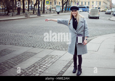 Les jeunes, de la hanche et attractive blonde hailing taxi , fille d'un joli chapeau et un manteau gris Banque D'Images