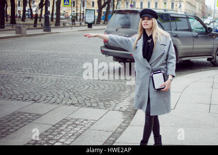 Les jeunes, de la hanche et attractive blonde hailing taxi , fille d'un joli chapeau et un manteau gris Banque D'Images