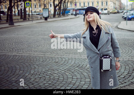 Les jeunes, de la hanche et attractive blonde hailing taxi , fille d'un joli chapeau et un manteau gris Banque D'Images