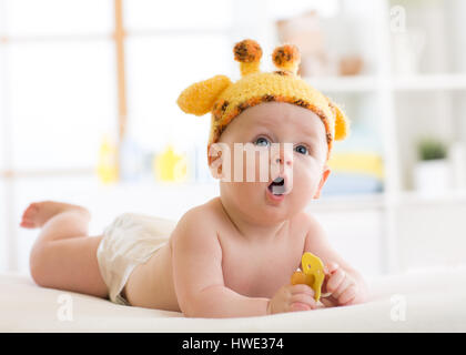 Funny baby boy in girafe hat allongé sur le ventre en pépinière. Petit enfant se trouve sur le lit avec bouche ouverte et est titulaire d'sucette dans les mains Banque D'Images