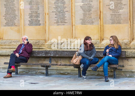 Cirencester - Trois personnes assis sur un banc - deux jeunes femmes bavardant tandis que man talks on mobile phone at Cirencester, Gloucestershire en Mars Banque D'Images