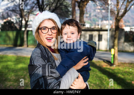 Portrait of young woman in santa hat carrying baby boy in park Banque D'Images