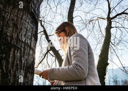 Jeune femme debout à côté arbre, lecture, livre, low angle view Banque D'Images