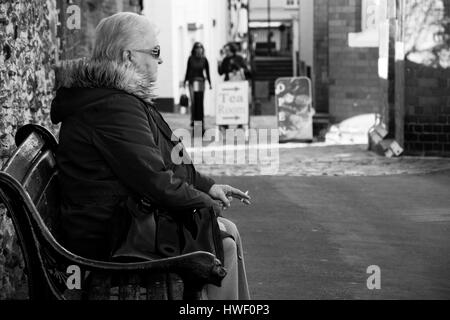 Une vieille femme assis sur un banc, fumant une cigarette. Banque D'Images