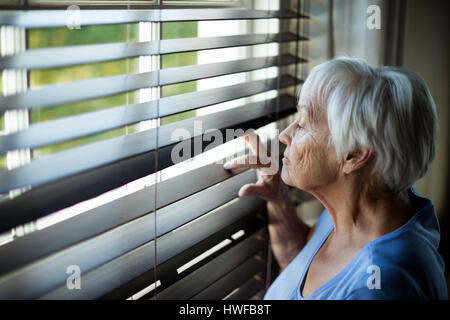 Senior woman looking out from window blind à la maison Banque D'Images