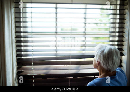 Senior woman looking out from window blind à la maison Banque D'Images