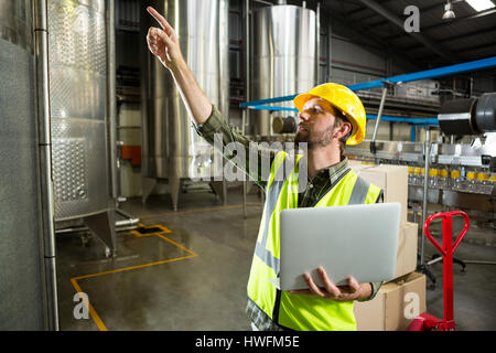 Travailleur homme pointe vers le haut tout en maintenant l'ordinateur portable en usine Banque D'Images