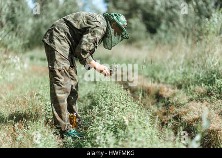 Femme en tenue de camouflage vêtements de protection les feuilles de menthe de collecte par les mains, avec la nature mise au point sélectionné forêt vert brillant, fond lumière ensoleillée Banque D'Images