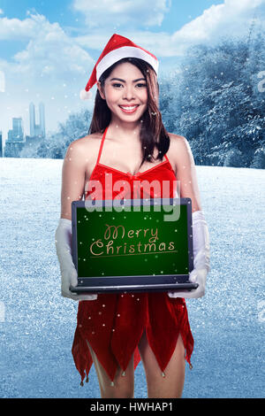 Asian woman wearing santa claus costume holding laptop avec joyeux noël écrit Banque D'Images