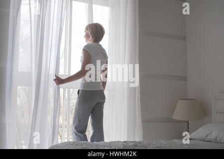 Jeune jolie fille d'ouvrir les rideaux le matin pour prendre l'air, vue de dos, concept blanc Banque D'Images