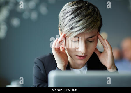 Portrait of young businesswoman with closed eyes souffrant de maux de tête alors que working on laptop in modern office Banque D'Images