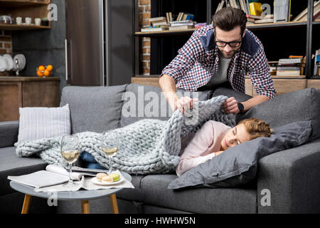 Jeune fille s'endormit sur le canapé et son petit ami qui couvre sa couverture de laine avec Banque D'Images