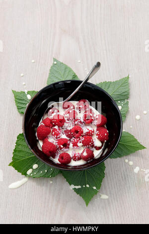 Framboises fraîches et crème en noir bol avec cuillère à thé et feuilles de framboisier, sur fond de similibois Banque D'Images