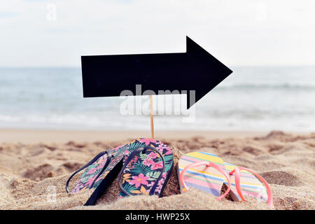 Libre d'une pancarte vide noir dans la forme d'une flèche signe et deux paires de tongs sur le sable d'une plage Banque D'Images