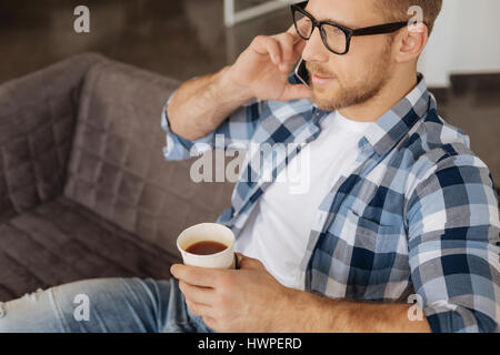 Avoir une pause. Smart nice homme barbu assis sur le canapé et parler au téléphone tout en prenant le thé Banque D'Images