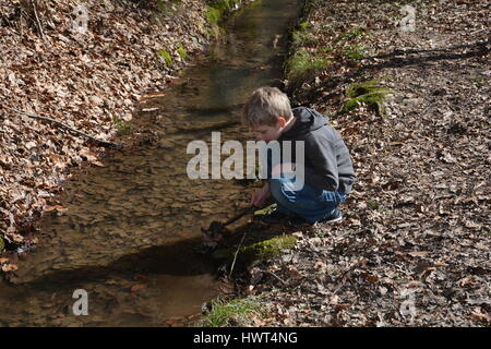 Petit garçon blond dans un petit ruisseau joue avec une branche dans l'eau Banque D'Images