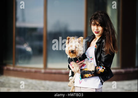 Brunette gitane avec yorkshire terrier dog posés contre de grandes fenêtres maison. L'usure du modèle sur blouson de cuir et t-shirt avec l'ornement, des pantalons. Banque D'Images