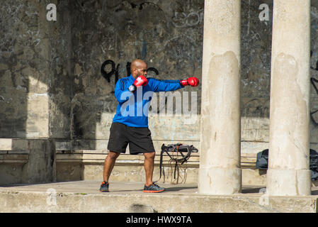 Un male boxer noir rouge vif avec des gants de boxe pratiquer la boxe à un bâtiment délabré avec piliers sur une journée ensoleillée. Banque D'Images