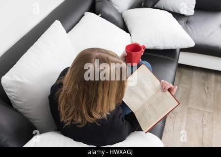 Vue aérienne de jeune femme lisant un livre sur un canapé noir et blanc noir et blanc avec des oreillers. Woman is holding a mug rouge dans sa main gauche. Le te Banque D'Images