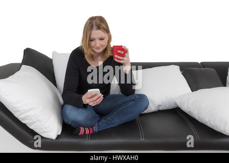 Jeune femme est à l'aide de son téléphone intelligent tenant une tasse rouge et assise en tailleur sur un canapé noir et blanc noir et blanc avec des oreillers. Isolé sur t Banque D'Images