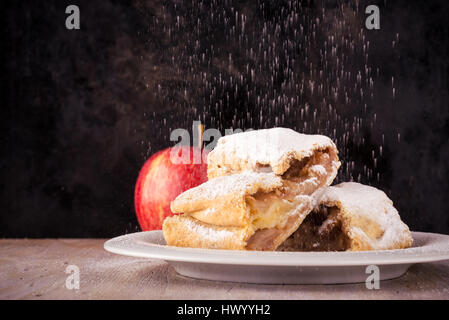 Photo horizontale avec deux tranches de strudel et seule pomme rouge. Deux portions sont couverts par du sucre en poudre qui est renversé par le haut sur une soucoupe blanche. Banque D'Images