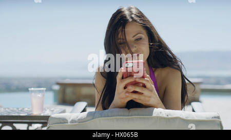 Close up of woman in bikini en utilisant un téléphone cellulaire et bronzer sur des chaises près de la piscine Banque D'Images