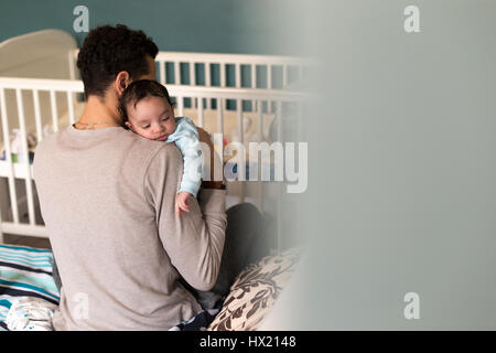 Jeune père est assis sur le bord de son lit avec son bébé blotti dans son épaule. Banque D'Images