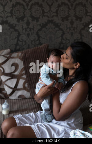 Jeune mère est assise sur son canapé et tenant son bébé qui vient d'être nourris. Elle est de l'embrasser sur la tête avec lui a tenu près d'elle. Banque D'Images